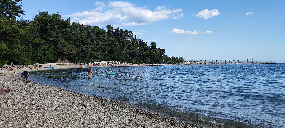 Pebble shore, swimmers and wooded hillside at Kašjuni Beach in Split